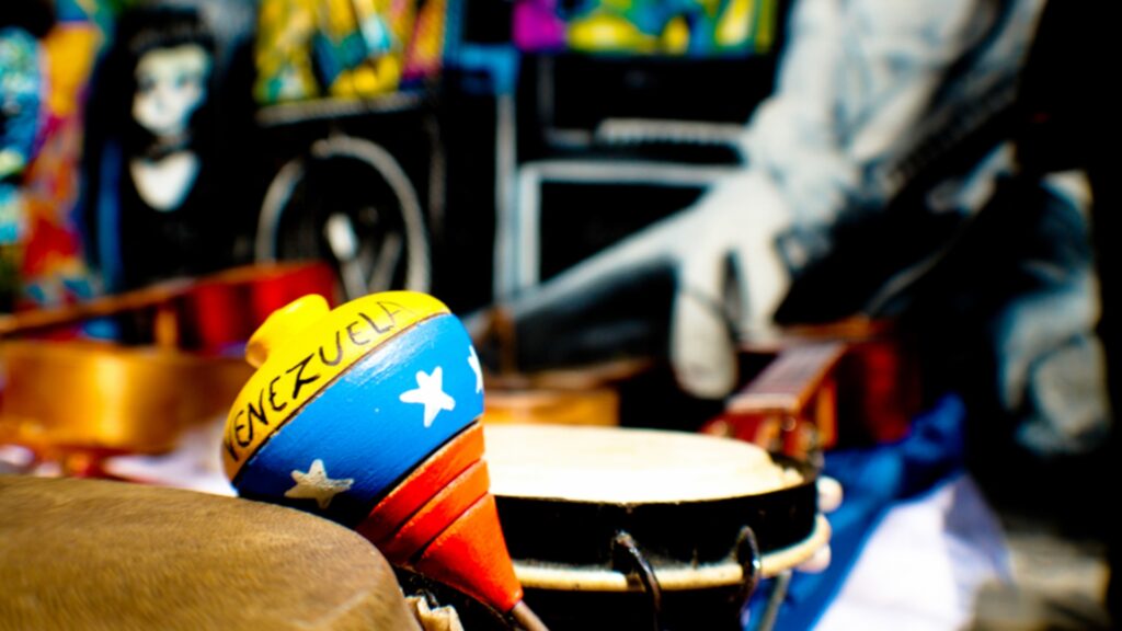 Various musical instruments blurred out with a colorful maraca in focus painted in the colors and starts of the Venezuelan flag with the word Venezuela written in black on yellow background.