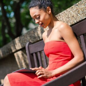 Hispanics and Latinos are often used incorrectly and are not interchangeable. Mixed race Latina sitting on a park bench wearing a red dress and using a tablet.