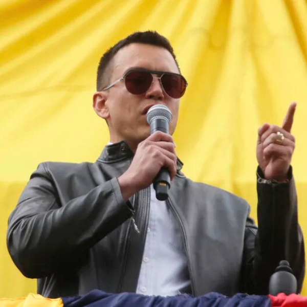 Ecuadorian President, Daniel Noboa, wearing jacket and sunglasses and holding microphone while giving speech with a yellow background.