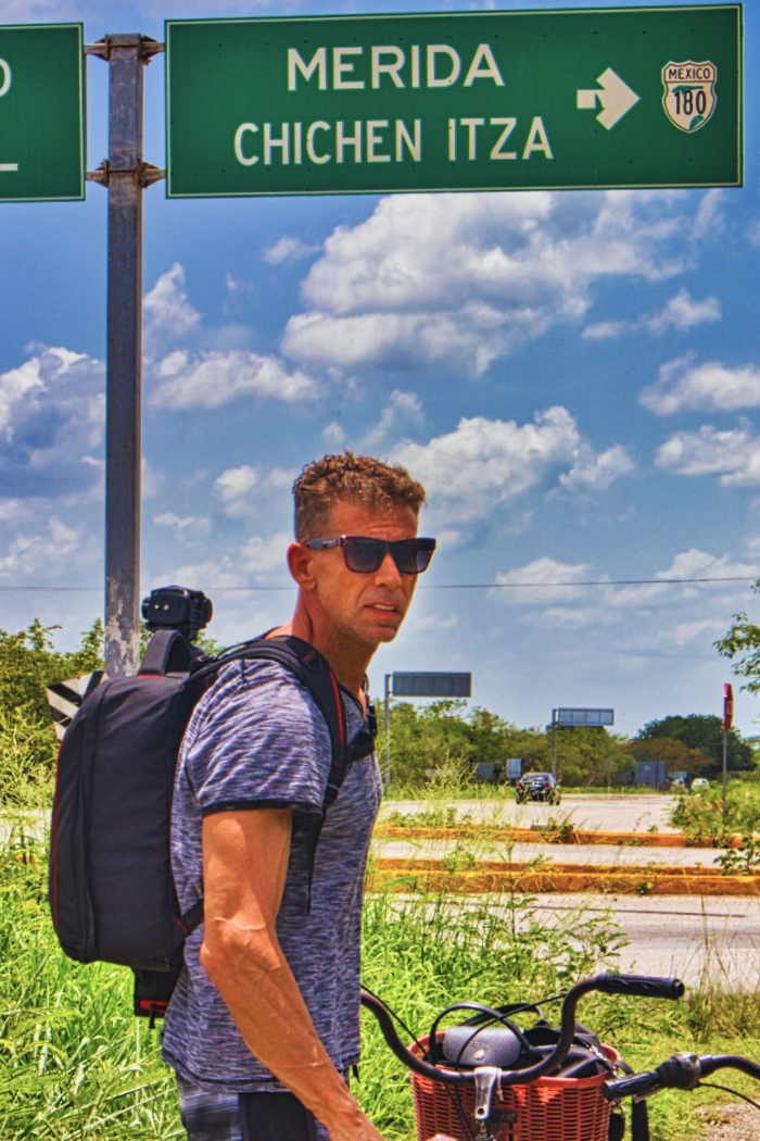 Eric standing to the side of a bicycle with grey t-shirt and black backpack in front of a road sign indicating direction to Mérida and Chichen Itza in Yucatán, Mexico.