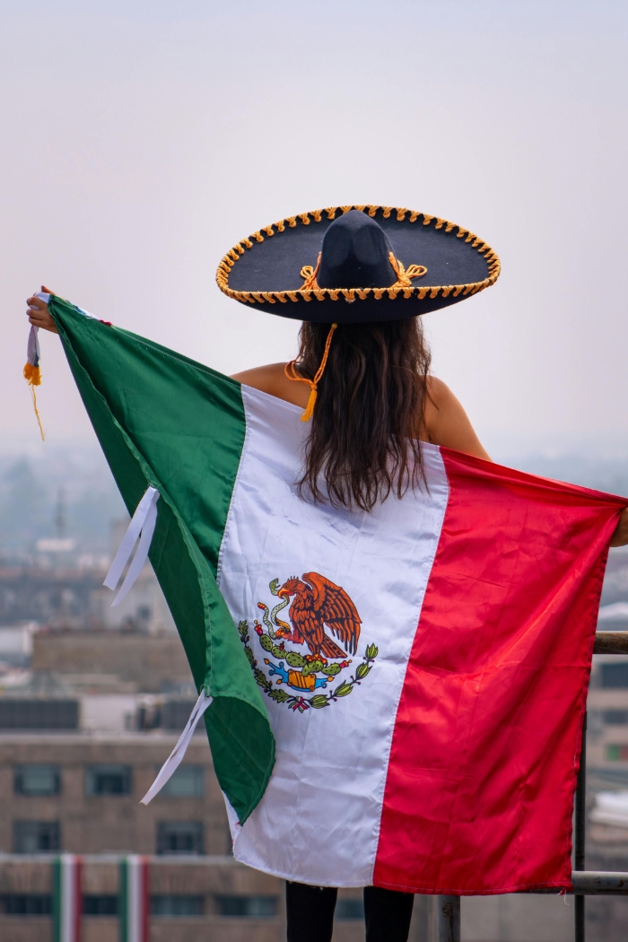 Mexican girl with black and gold sombrero with back turned, holding the Mexican Flag.