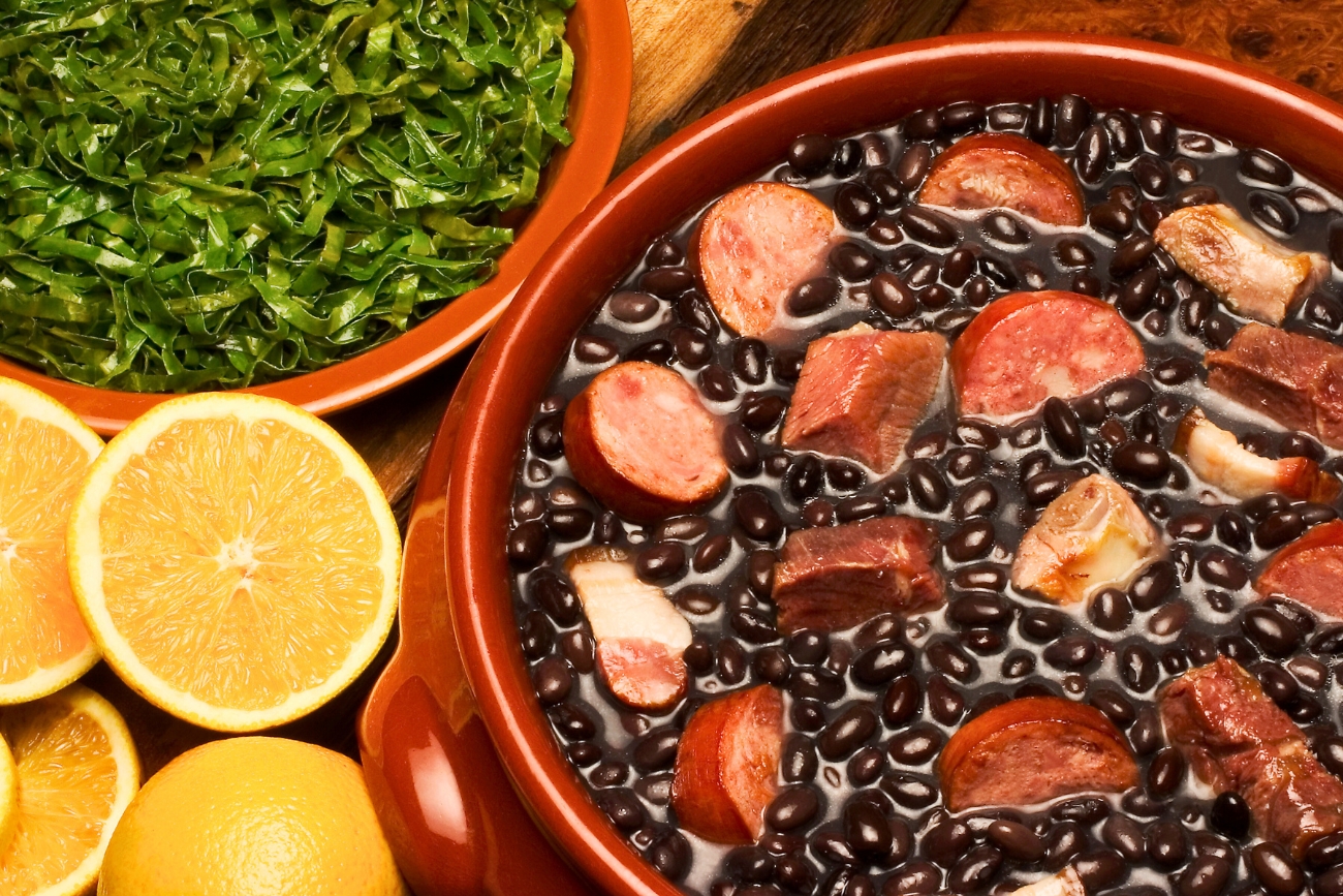 Feijoada, Brazil's hearty meat and bean national dish in a pot on a table next to sliced lemons and leafy greens.