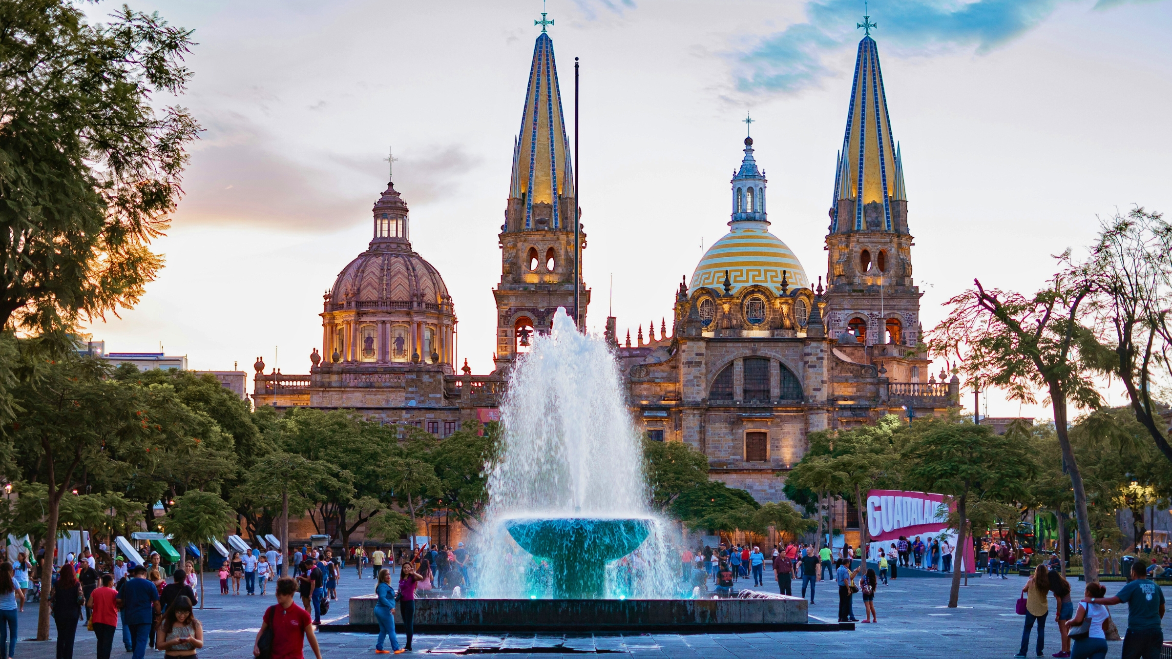 The sand, yellow and blue colored cathedral called Catedral basílica de la asunción de María santísima at dusk behind a sprouting water fountain with lots of people out and about in Guadalajara, Jalisco, Mexico.