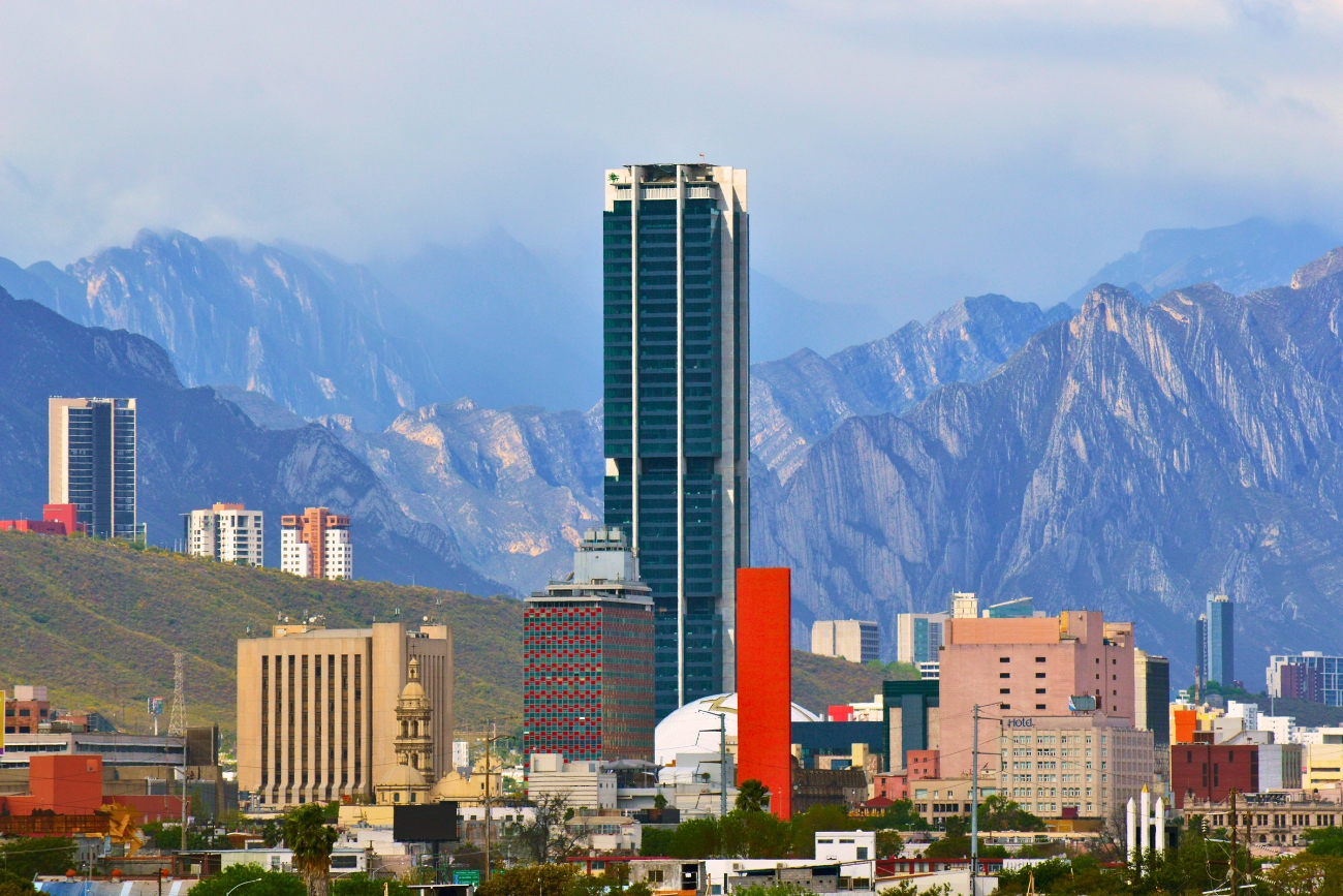 Cityscape showing 1 tall skyscraper and numerous buildings with impressive mountains and cloud cover in the background in Monterrey, Nuevo León, Mexico.