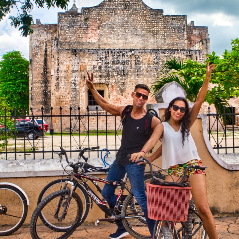 Dani and Eric sitting on bikes waving to the camera in Valladolid, Yucatán, Mexico.