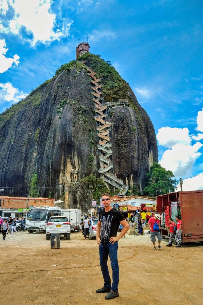 Eric wearing sunglasses on a partly cloudy day, standing in front of the monolith, El Peñol de Guatapé in Antioquia, Colombia.