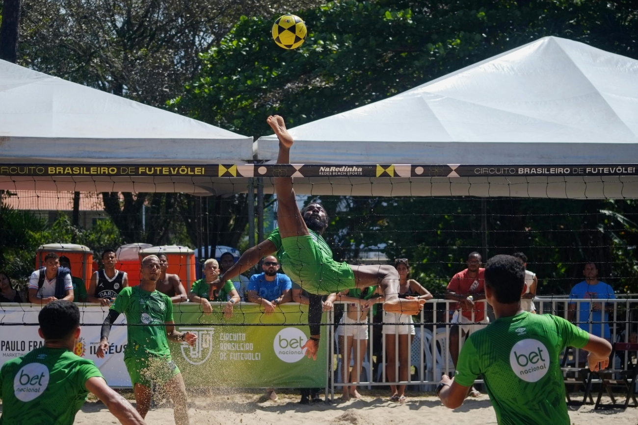 A player for Bet Nacional is positioned upside down as his leg and foot go high above the net to strike the ball in a Futevôlei tournament in Brazil.