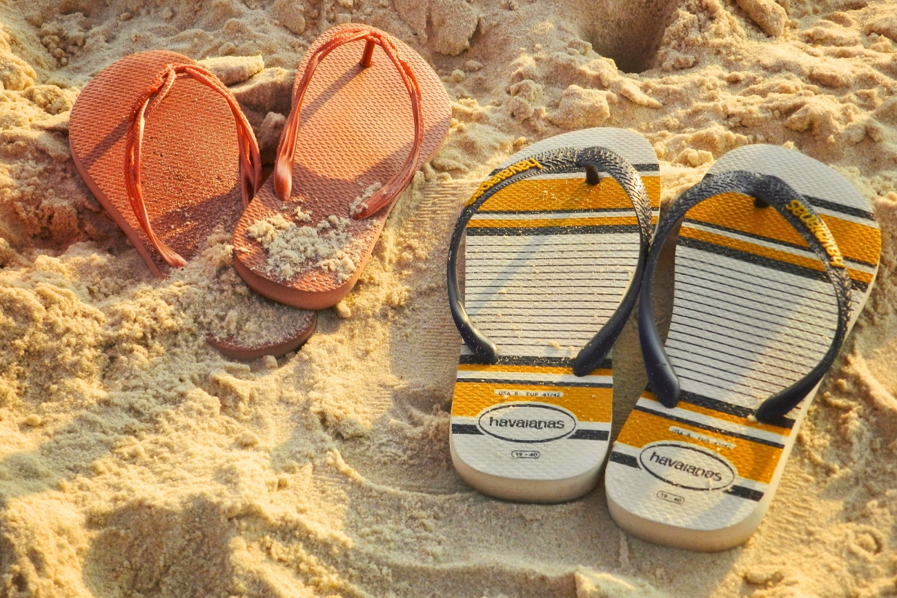 Two pairs of Brazilian Havaianas, coral and off white with yellow and black bars lying in the sand.