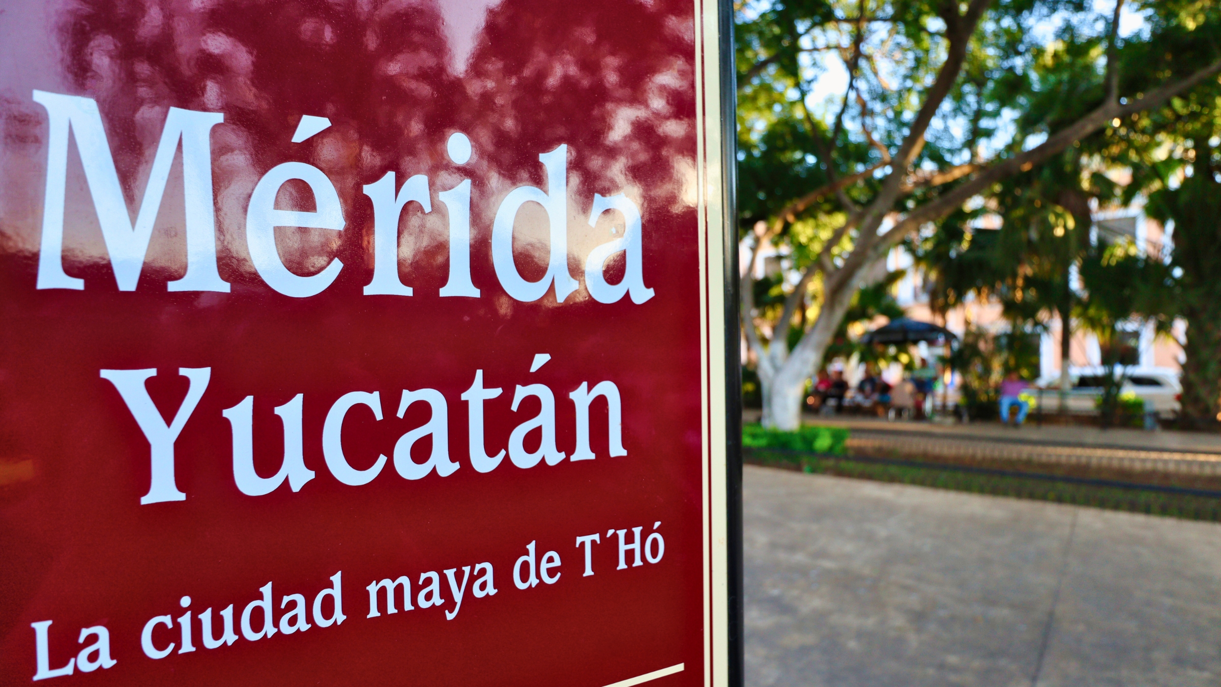 A sign of that reads "Mérida Yucatán, la cuidad maya de T'Hó" in white with maroon colored background with lush vegetation and people shown in bokeh to the right of the sign.