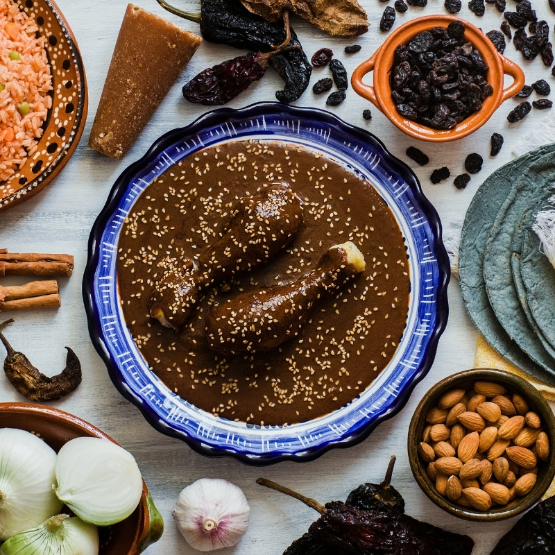 Chicken legs in a bowl with brown Oaxacan mole along with rice, chiles, raisins, nuts, onions and garlic around the bowl.