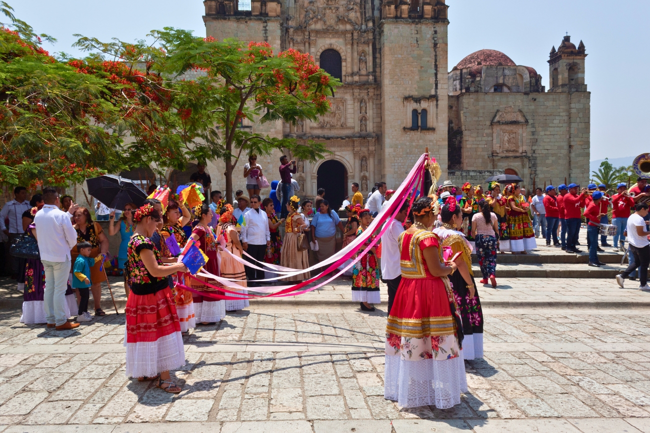 Parade or ceremony with women dressed in coloful outfits and a male band dressed in red and blue playing instruments in the middle right, in front of a cathedral in Oaxaca, Oax, Mexico.