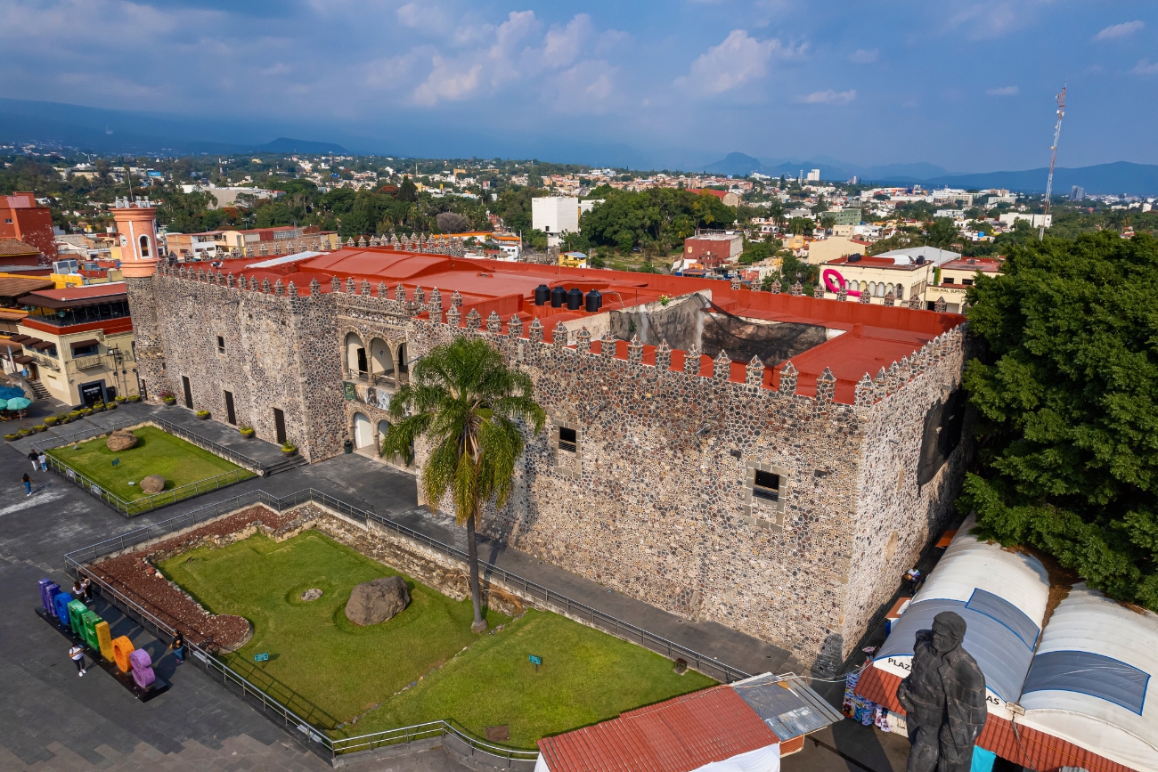Aerial view of Palacio Cortés with a solo palm tree and male statue on the bottom right on a partly cloudy day in Cuernavaca, Moreles, Mexico.