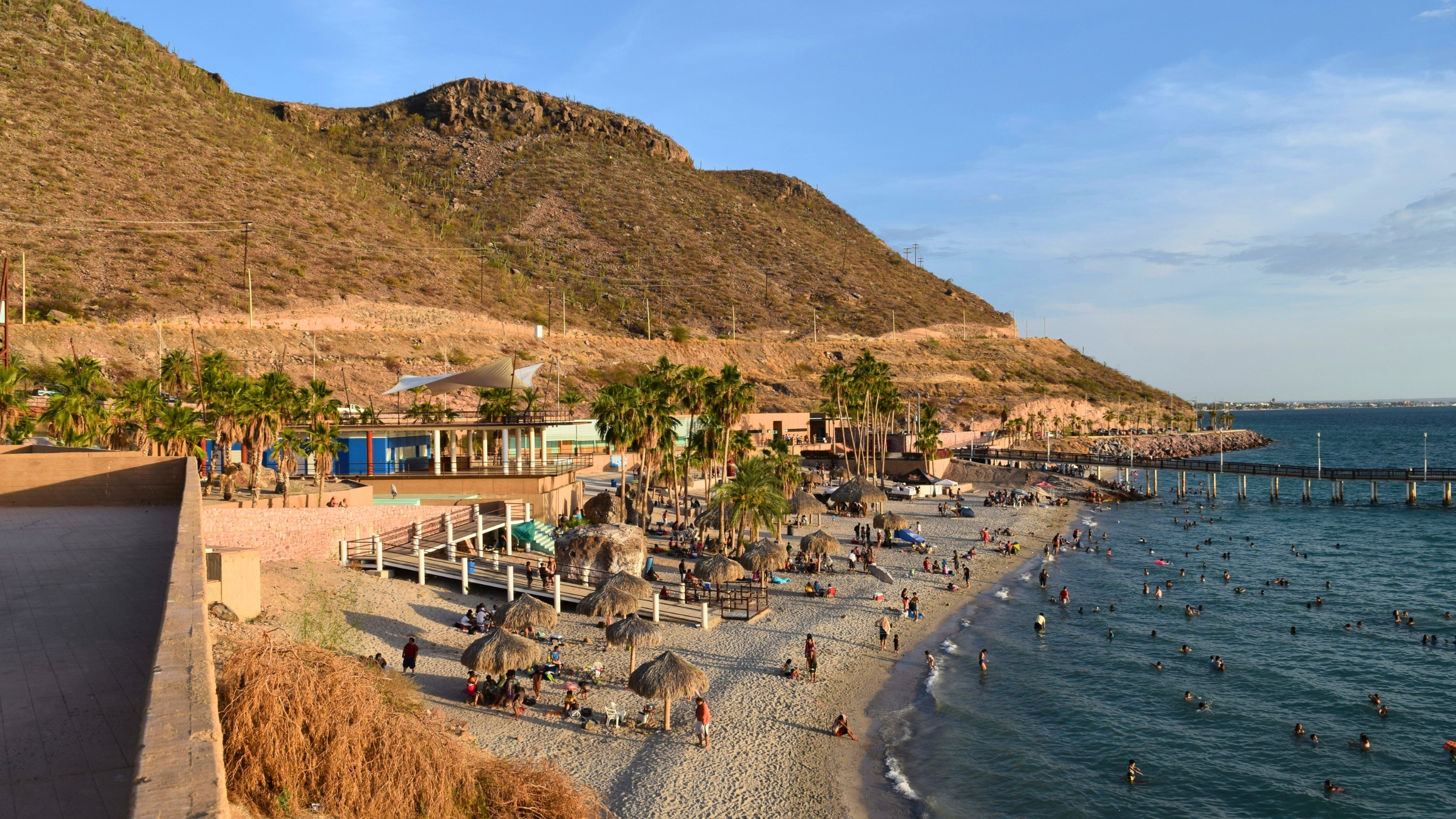 Playa Coromuel on a clear day showing lots of bathers in the water, palapas, palm trees, a pier and mountains in the background.