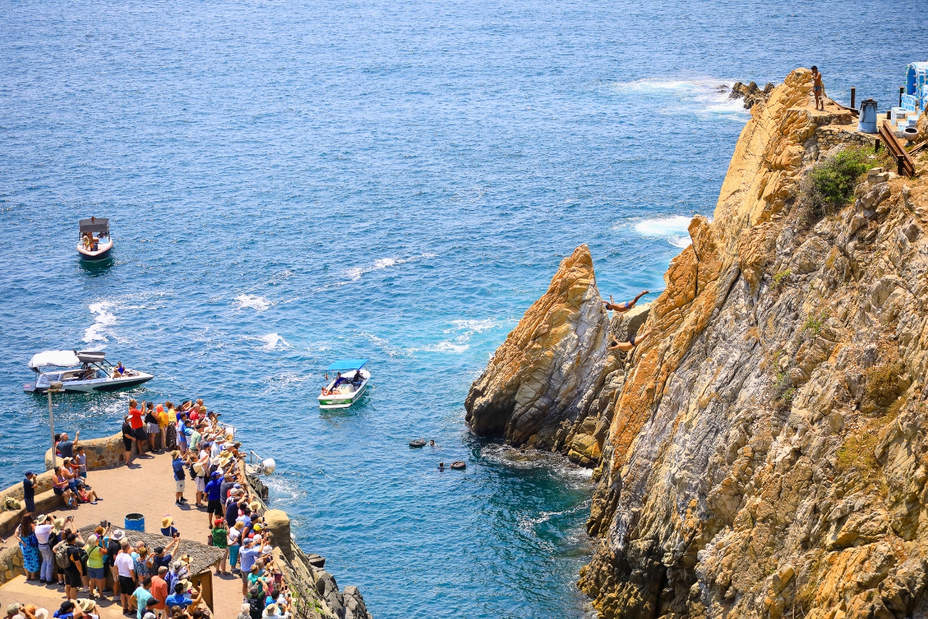 2 La Quebrada cliff divers in mid-flight, one diver waiting at the top with plenty of onlookers and 3 small boats in the background on a sunny day in Acapulco, Guerrero, Mexico.