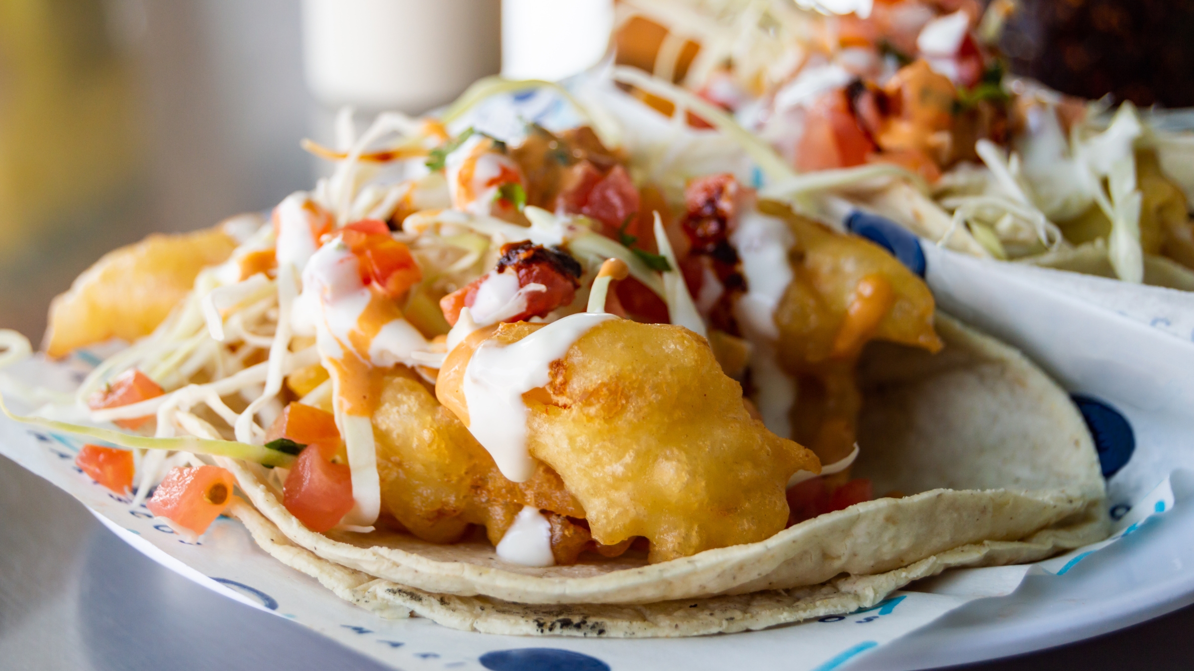 Scrumptious looking tempura style seafood tacos served on a white flour tortilla topped with coleslaw, diced tomatoes and sour cream.