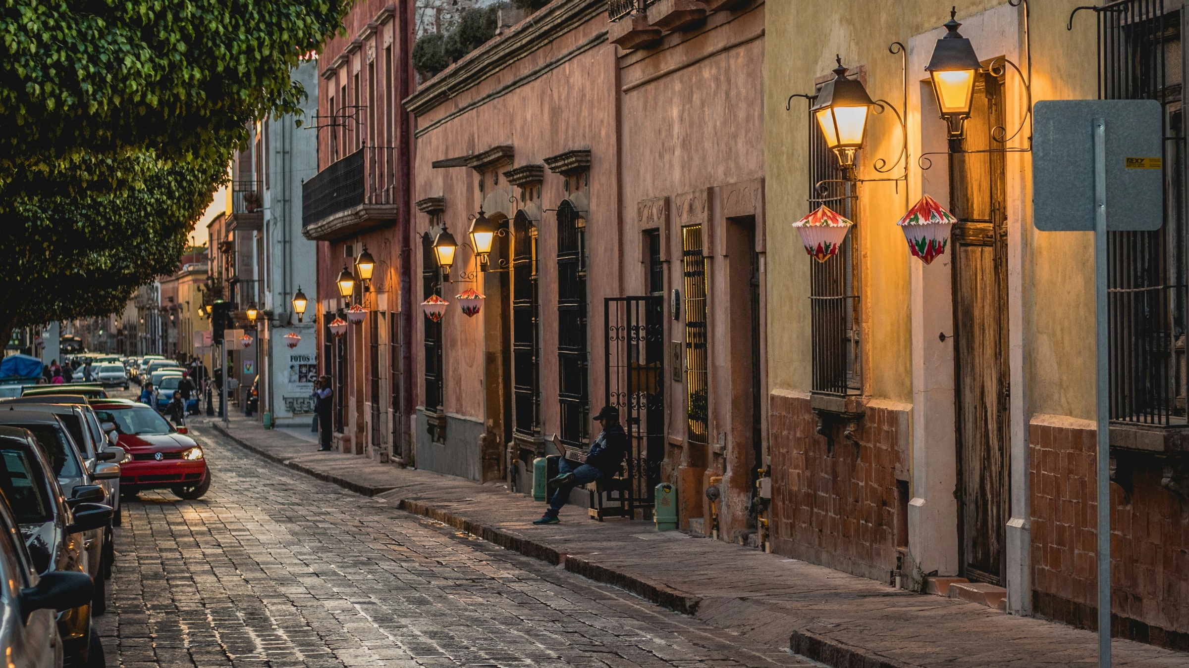 A street scene at dusk with street lights on showing a man sitting on a bench and a man standing on a sidewalk as a red Volkswagen, pulls out onto the stone paved street in Querétaro, Qro, Mexico.