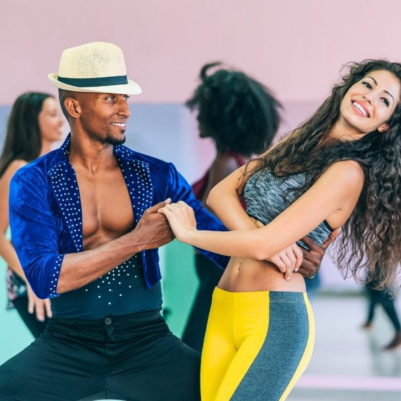 White woman tummy in outfit and dark-skinned man wearing white hat and open shirt dancing salsa with 2 female dancers in the background.