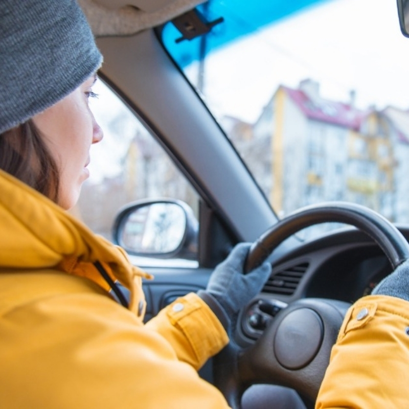 A woman in cold weather driving a car wearing a yellow jacket, grey hat and gloves.