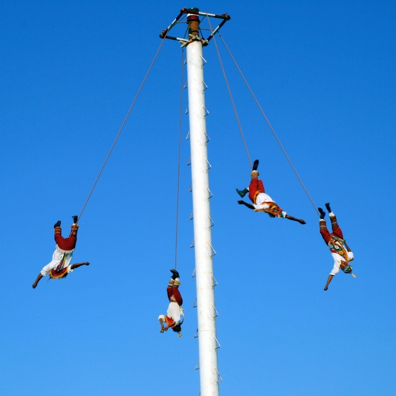 4 Voladores de Papantla from Veracruz, Mexico seen flying around white pole with rope attached to one leg on a clear day.