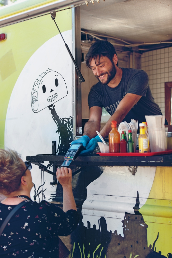 A bearded man working as a taco vendor holds a mobile payment terminal for a woman to pay at a taco stand.